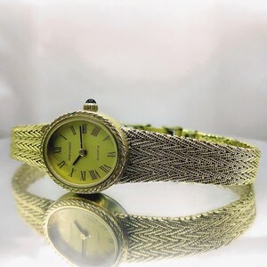 May include: A gold-toned Movado quartz watch with a woven strap. The watch face is round with Roman numerals and a light green background. The watch is reflected in the shiny surface below.