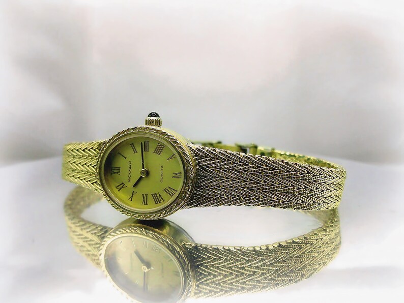 May include: A gold-toned Movado quartz watch with a woven strap. The watch face is round with Roman numerals and a light green background. The watch is reflected in the shiny surface below.