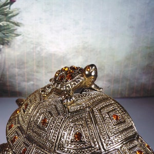 May include: A gold-toned turtle trinket box with a smaller turtle on top. The turtle is decorated with amber-colored rhinestones.
