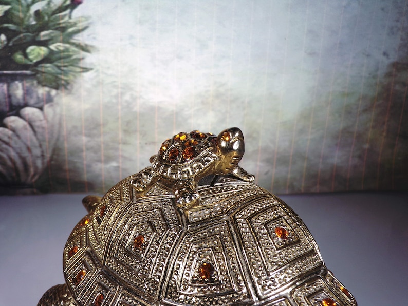 May include: A gold-toned turtle trinket box with a smaller turtle on top. The turtle is decorated with amber-colored rhinestones.