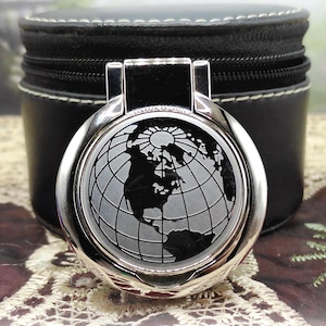 Può includere: Un specchietto compatto rotondo color argento con un design dettagliato della mappa del mondo. Lo specchio è posto contro un astuccio in pelle nera con chiusura a cerniera. Il design presenta un sole e i continenti.