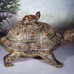 May include: A gold-toned turtle trinket box with a smaller turtle on top. The turtle is decorated with orange rhinestones.