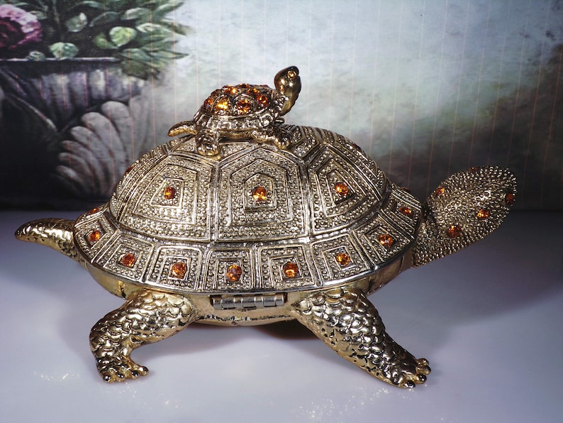 May include: A gold-toned turtle trinket box with a smaller turtle on top. The turtle is decorated with orange rhinestones.