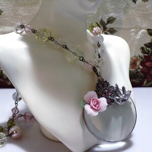 Peut inclure: Un collier d&eacute;licat avec un pendentif en forme de loupe. Le pendentif pr&eacute;sente une fleur rose et un motif d'oiseau avec des strass. Le collier est compos&eacute; de perles de verre transparentes, roses et vertes.