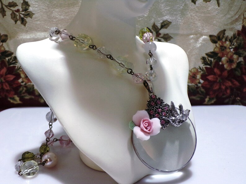 Peut inclure: Un collier d&eacute;licat avec un pendentif en forme de loupe. Le pendentif pr&eacute;sente une fleur rose et un motif d'oiseau avec des strass. Le collier est compos&eacute; de perles de verre transparentes, roses et vertes.