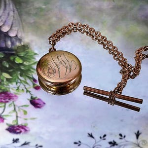 May include: A gold-toned pocket watch with an engraved design, attached to a chain and a T-bar. The watch is round with a hinged lid. The chain and T-bar are also gold-toned. The background has floral and botanical elements.