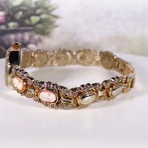 May include: Gold-toned bracelet watch with oval cameo designs and a decorative link band. The watch features a small, round face and is set against a soft, blurred background.