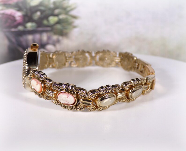 May include: Gold-toned bracelet watch with oval cameo designs and a decorative link band. The watch features a small, round face and is set against a soft, blurred background.