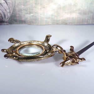 May include: Gold-toned turtle-shaped magnifying glass with a clear glass lens. The magnifying glass is separated into two parts, the base and the handle. The handle is a smaller turtle-shaped piece with a thin metal rod extending from its back.