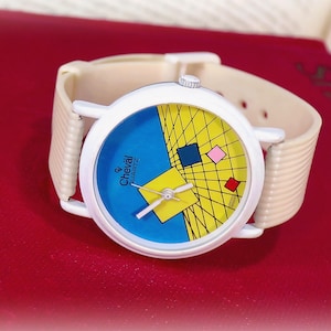 May include: A wristwatch with a white round case and a beige ribbed strap. The watch face features a blue and yellow geometric design with a grid pattern, small colored squares, and the text Cheval Quartz printed in black on the left side.
