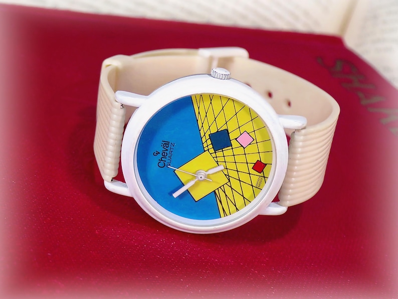 May include: A wristwatch with a white round case and a beige ribbed strap. The watch face features a blue and yellow geometric design with a grid pattern, small colored squares, and the text Cheval Quartz printed in black on the left side.