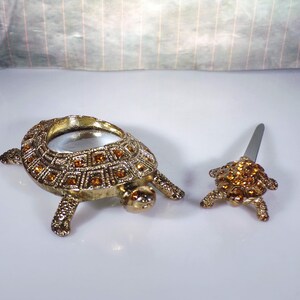 May include: Two gold turtle figurines with rhinestones. The larger turtle has a magnifying glass in its shell. The smaller turtle has a small knife in its shell.