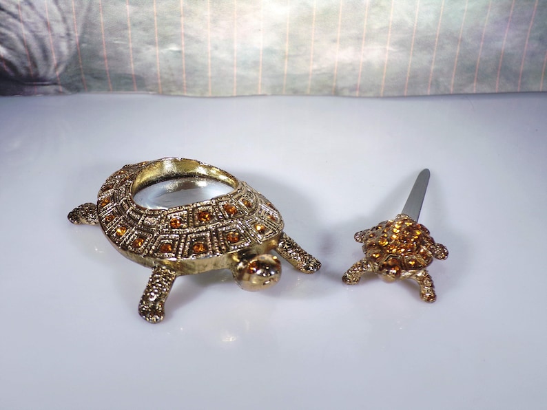 May include: Two gold turtle figurines with rhinestones. The larger turtle has a magnifying glass in its shell. The smaller turtle has a small knife in its shell.