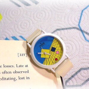 May include: A white analog wristwatch with a round face featuring a blue background, a yellow grid pattern, and small red, blue, and pink squares. The watch has a white case and a textured, off-white ribbed strap, with the brand name Cheval Quartz.