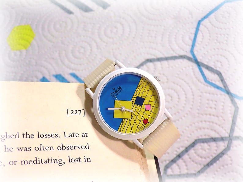May include: A white analog wristwatch with a round face featuring a blue background, a yellow grid pattern, and small red, blue, and pink squares. The watch has a white case and a textured, off-white ribbed strap, with the brand name Cheval Quartz.