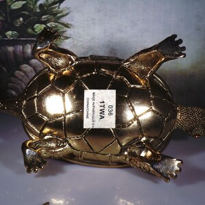 May include: A gold-colored turtle figurine with a detailed shell and legs. The turtle is lying on its back with its legs outstretched. The figurine has a label on its shell that reads "036 1TTWA MADE IN/FABRIQU&Eacute; EN CHINACHINE".