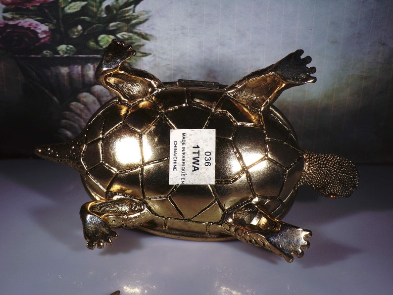 May include: A gold-colored turtle figurine with a detailed shell and legs. The turtle is lying on its back with its legs outstretched. The figurine has a label on its shell that reads "036 1TTWA MADE IN/FABRIQU&Eacute; EN CHINACHINE".
