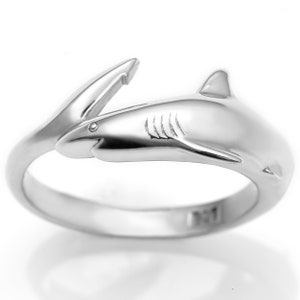 May include: A silver shark ring. The ring showcases a detailed shark design, including a dorsal fin, gills, and a pointed snout. The shark's body curves around the finger, creating a unique piece of jewellery.