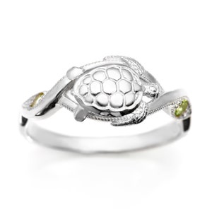 May include: A silver ring with a detailed turtle design and two green gemstones. The ring is a twisted band with a smooth finish.