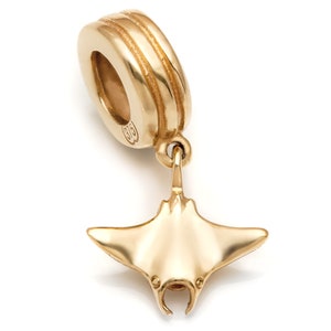 Puede incluir: Un charm dorado con un diseño de manta raya. El charm tiene un lazo en la parte superior para sujetarlo a una pulsera o collar. La manta raya tiene una superficie detallada y texturizada y está suspendida del lazo.
