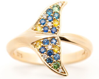 May include: A gold-colored ring featuring a whale tail design. The tail is adorned with a mosaic of blue, yellow, green, and white gemstones. The ring's band is smooth and polished, creating a striking contrast with the colorful tail.