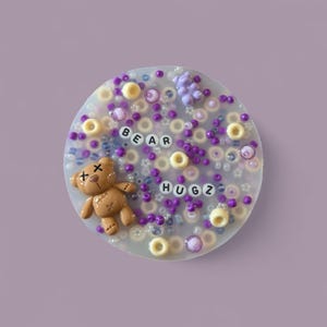 Op de afbeelding: Een ronde, witte telefoonhouder van hars met een bruine teddybeer met een 'X' op zijn ogen, paarse kralen en de woorden 'BEAR HUGZ' in paarse letters.