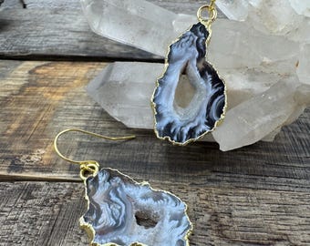 Puede incluir: Un par de pendientes de ágata con borde dorado. Los pendientes presentan una forma natural e irregular con tonos grises, blancos y negros. Los pendientes se exhiben sobre una superficie de madera.