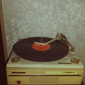 May include: A vintage record player with a black vinyl record on the turntable. The record player is white with a brown trim and has a silver tone arm. The record player has a label on the front that says "3350 E".
