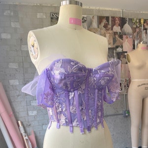 Pu&ograve; includere: Un top a corset in pizzo floreale viola e bianco con un'overlay trasparente e spalline regolabili. Il top &egrave; su un manichino.