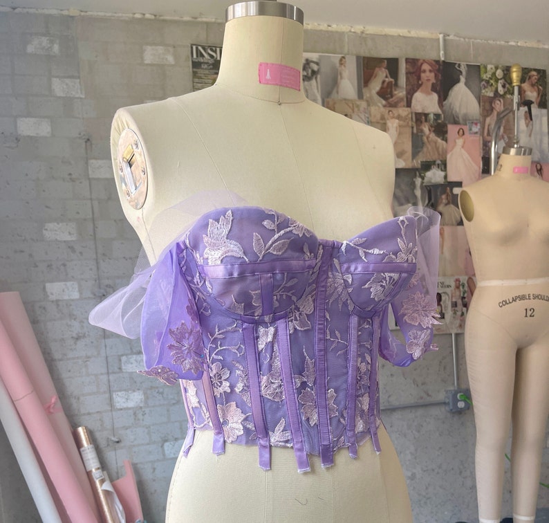 Pu&ograve; includere: Un top a corset in pizzo floreale viola e bianco con un'overlay trasparente e spalline regolabili. Il top &egrave; su un manichino.