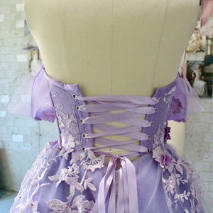 Pu&ograve; includere: Un abito lavanda senza spalline con un'applicazione di pizzo e una schiena a corsetto con un fiocco di nastro di raso. L'abito ha una gonna di tulle e ricami floreali.