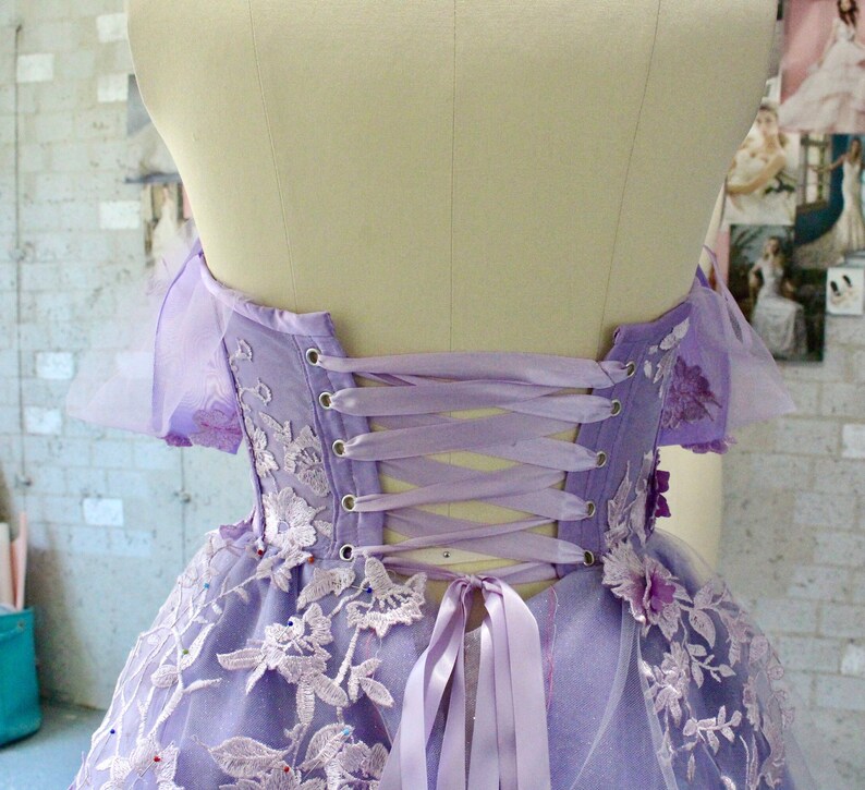 Pu&ograve; includere: Un abito lavanda senza spalline con un'applicazione di pizzo e una schiena a corsetto con un fiocco di nastro di raso. L'abito ha una gonna di tulle e ricami floreali.