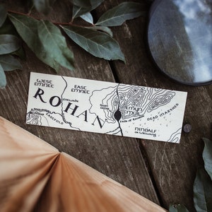 May include: A map of Rohan, with the text "ROHAN" prominently displayed. The map features detailed topographical lines and place names like "Dead Marshes" and "Falls of Rauros." The map is on a wooden surface with leaves and a magnifying glass.
