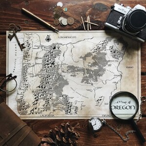 May include: A vintage-style map of Oregon with a magnifying glass on top. The map is printed on paper with a cream background and black text. The magnifying glass is round with a black frame and a clear lens. The text on the magnifying glass reads "Map of Oregon".