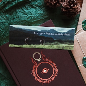 May include: A brown hardback book with a red and gold design of an eye and a ring on the front cover. The book is sitting on a green surface with a bookmark that says "Courage is found in unlikely places."