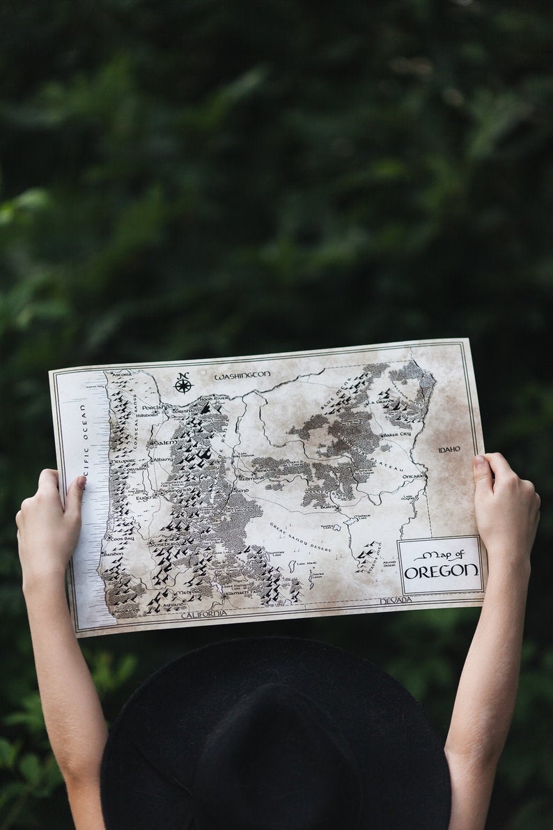 May include: A person holds a vintage-style map of Oregon, with the text "Map of Oregon" in a box at the bottom right corner. The map is printed on aged paper and shows the state's major cities, rivers, and mountains.