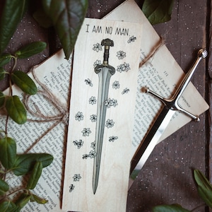 Puede incluir: Un marcapáginas de madera con una ilustración de una espada y el texto "I AM NO MAN." La espada está adornada con pequeños dibujos de flores. También se ve un abrecartas de metal y páginas de libro.