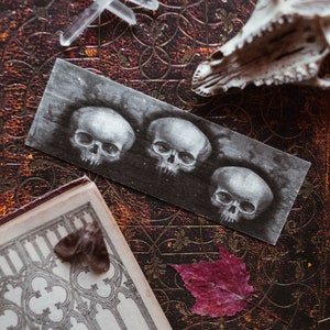 May include: A vintage book with a dark red leather cover and intricate gold designs. A piece of paper with three black and white skull drawings is placed on the book. A small white deer skull is on the top right corner of the book.