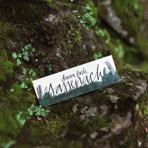 May include: A white card with green and black lettering that says "dinna fash.  Sassenach." The card is lying on a bed of green moss.