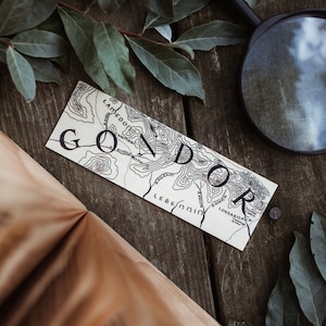 Könnte beinhalten: Eine Nahaufnahme einer Karte von Gondor aus Der Herr der Ringe. Die Karte ist auf weißem Hintergrund mit schwarzen Linien und Text gedruckt. Die Karte liegt auf einer Holzoberfläche mit grünen Blättern.