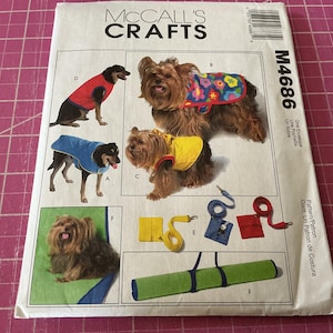 May include: A McCall's Crafts M4686 sewing pattern envelope featuring illustrations of small dogs wearing various colored coats, including red, blue, yellow, and floral patterns, along with matching leashes, small pouches, and a green mat.