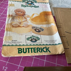 May include: A Butterick sewing pattern envelope for Cabbage Patch Kids doll clothing. The white paper envelope features a graphic of a doll in a yellow outfit, a yellow basket, and the text Cabbage Patch Kids, P'tits Bouts de Choux, and Butterick.