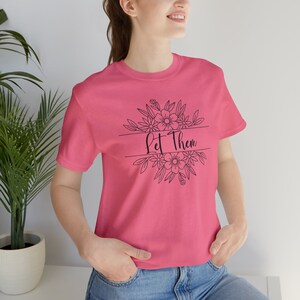 Pode incluir: Camiseta rosa com um design floral preto e o texto "Let Them" em um círculo.