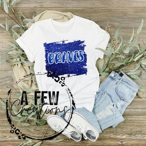 May include: White t-shirt with the word "BRAVES" in blue lettering on a blue glitter background. The shirt is paired with light blue denim shorts and white sneakers. The image is styled on a wooden surface with greenery.