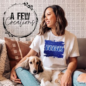 May include: A woman wearing a white t-shirt with a blue glitter design that says "BRAVES" on it. She is sitting on a sofa with a brown and white patterned cushion and a small brown and white dog.
