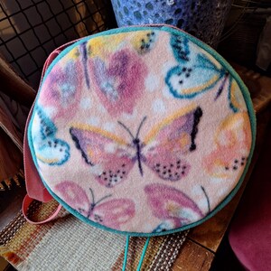 May include: A round, soft-textured crossbody bag featuring a light pink background with a repeating pattern of butterflies in shades of pink, yellow, and blue. The bag has a teal-colored trim around the circular edge and a pink strap.