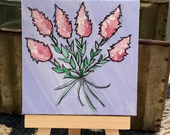 May include: A small painting on a wooden easel depicts a bouquet of pink flowers with green leaves on a purple background.