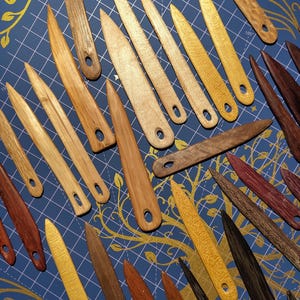 May include: A selection of wooden tools, possibly for crafts or bookbinding, in shades of brown and yellow. Each tool has a pointed end and a small hole. The tools are displayed on a blue surface with a gold pattern.