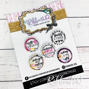 Puede incluir: Cinco botones redondos de 2,5 cm de diámetro con diseños de maquillaje. Los botones son blancos con detalles negros y rosas. El texto en los botones dice: "Hello Gorgeous", "Makeup Artist in Training", "Makeup is my Art", "Wake Up and Makeup" y "Flawless & Fierce".
