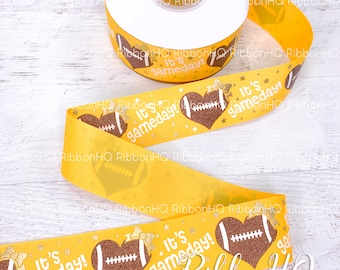 May include: Yellow ribbon with a white background and gold glitter. The ribbon has a repeated pattern of a brown football in a heart shape with a white bow and the text "It's gameday!"